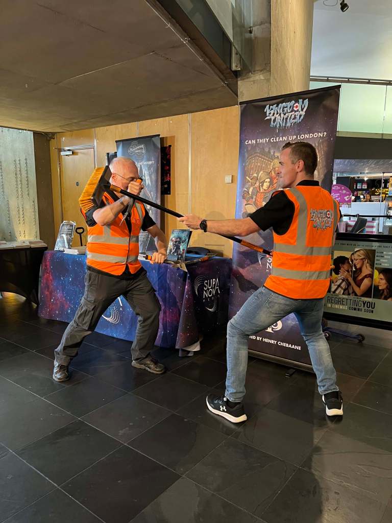 Martial artists Pete Halloran and Matt Law, as Supanova Media stand during 2025 Fighting Spirit Film Festival in Hackney. Wearing high-viz vest and sparring with cleaning tools from the graphic novel Kingdom United by Henry Chebaane.